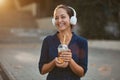 Cheerful laughing female holding cup of drink. Woman wearing white headset outdoors. Take break and coffee time concept Royalty Free Stock Photo