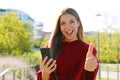 Cheerful laughing young woman with mobile phone giving thumb up outside Royalty Free Stock Photo