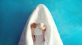 Cheerful lazy woman waking up after sleeping covered head with white soft blanket holds cup of coffee on blue background at home Royalty Free Stock Photo