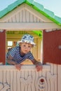 A cheerful little boy in a hat, inside a toy little house, he looks out the window with the shutters straight into the camera Royalty Free Stock Photo