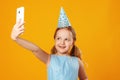 Cheerful little girl celebrates birthday. The child holds the phone, takes a selfie. Closeup portrait on yellow background