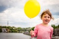 A cheerful little girl is holding a yellow ball in her hand. The child stands on the background of the river and city park. Happy Royalty Free Stock Photo