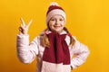 Cheerful little girl in a jacket, scarf and hat on a yellow background. The child shows a victory sign. Success concept Royalty Free Stock Photo