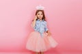 Cheerful little girl with long brunette hair in tulle skirt holding princess crown on head isolated on pink background Royalty Free Stock Photo