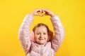 Cheerful little girl shows a heart with her fingers over her head. A child in a jacket and warm ear muffs on a yellow background. Royalty Free Stock Photo