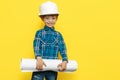 Cheerful little kid boy engineer or architect in a protective helmet holding construction plan over yellow background. Royalty Free Stock Photo