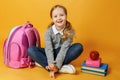 Cheerful little student girl is sitting with backpack, books and lunch box on yellow background. Child student looks into the Royalty Free Stock Photo