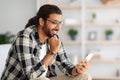 Cheerful long-haired middle-eastern guy hipster sitting on couch, using smartphone Royalty Free Stock Photo