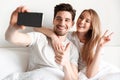 Cheerful loving couple lies in bed take a selfie showing peace gesture. Royalty Free Stock Photo