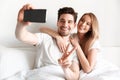 Cheerful loving couple lies in bed take a selfie showing peace gesture. Royalty Free Stock Photo