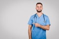 Cheerful male doctor in blue, pointing to side at blank space on grey background