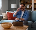 Cheerful male freelancer talking over mobile phone and working over laptop on table while sitting on sofa. Young businessman Royalty Free Stock Photo