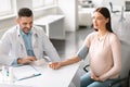 Cheerful male gynecologist measuring blood pressure for young pregnant patient during checkup in clinic