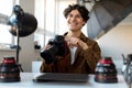 Cheerful male photographer sitting at workplace in modern photostudio, holding professional camera and smiling Royalty Free Stock Photo