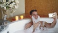 Cheerful man with foam on beard is taking selfie using smartphone in hot tub in modern spa salon. He is laughing