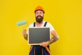 cheerful man housepainter in helmet and work clothes hold paint roller and blackboard