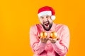 A cheerful man in a Santa Claus hat on a Christmas yellow background holds in front of him balls of golden color on the Christmas Royalty Free Stock Photo