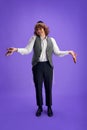 Cheerful man in tweed waistcoat and kippah with arms outstretched, holding grogger against purple studio background.