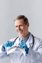 Cheerful and mature doctor in latex gloves tearing paper with coronavirus lettering