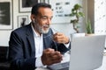 Cheerful mature european man in suit and headphones working on laptop in modern office interior Royalty Free Stock Photo