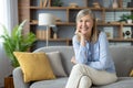 Cheerful mature woman sitting on decorated sofa with crossed legs and holding chin with palm. Positive female in casual Royalty Free Stock Photo