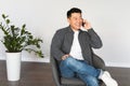 Cheerful middle aged asian man calling by phone, sitting in armchair in living room interior with white wall Royalty Free Stock Photo