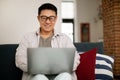 Cheerful middle aged asian man using laptop while resting on sofa, surfing internet or reading good news and smiling Royalty Free Stock Photo