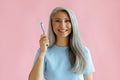 Cheerful middle aged Asian woman holds toothbrush standing on pink background