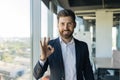 Cheerful middle aged businessman in formal wear showing ok sign and smiling at camera, standing in office interior Royalty Free Stock Photo