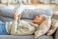Senior woman reading book and smiling while relaxing on sofa at home Royalty Free Stock Photo