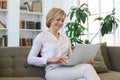 Cheerful middle aged woman using laptop while sitting on sofa at home