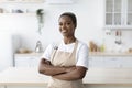 Cheerful millennial african american female in apron with crossed arms looking at camera at modern minimalist kitchen Royalty Free Stock Photo