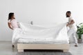 Cheerful millennial african american husband and wife make white bed with blanket in bedroom interior