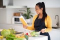 Cheerful millennial african american woman in apron reads new recipe in notebook or book, make salad Royalty Free Stock Photo