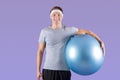 Cheerful millennial athlete with fitness ball smiling and looking at camera on lilac studio background