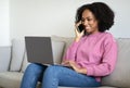 Cheerful millennial black woman typing on computer, reads message, calls by phone, sits on sofa Royalty Free Stock Photo