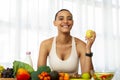 Cheerful millennial latin lady in sportswear eat green apple in light kitchen interior