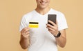 Cheerful millennial man in white t-shirt looks at smartphone and holds credit card, pays for purchase