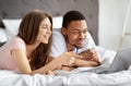 Cheerful millennial multinational couple lying on bed with laptop, watching fun video together Royalty Free Stock Photo