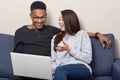 Cheerful mixed race couple use laptop computer for watching movies, sit together on sofa over home interior, browse information, Royalty Free Stock Photo