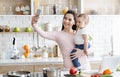 Cheerful mom with baby taking selfie on cellphone at kitchen Royalty Free Stock Photo