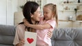 Cheerful mom embracing kid daughter holding postcard with red heart