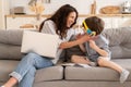 Cheerful mom and son laugh together on sofa: mother work on laptop and kid listen to audio lesson
