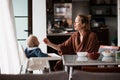 Cheerful mother wearing bathrope spoon feeding her infant baby boy child sitting in high chair at the dining table in Royalty Free Stock Photo