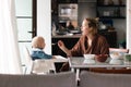Cheerful mother wearing bathrope spoon feeding her infant baby boy child sitting in high chair at the dining table in Royalty Free Stock Photo