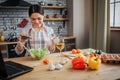 Cheerful nice woman sit at table in kitchen and put spices in bowl with salad. She looks happy. Woman wears apron