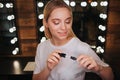 Cheerful nice young woman hold lipgloss and look on it. She stand in beauty room. Mirror with light bulbs behind. Royalty Free Stock Photo