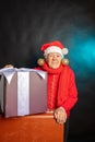 A cheerful, old woman, in a New Year`s cap, with a large present in her hands. Studio photo. Dark background