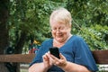 Cheerful old woman with smartphone at park. Outdoor portrait of happy senior woman at park using mobile phone. Plus size blond