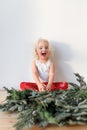 Cheerful overjoyed blonde female child sits crossed legs and Christmas garland in foreground, isolated over pink